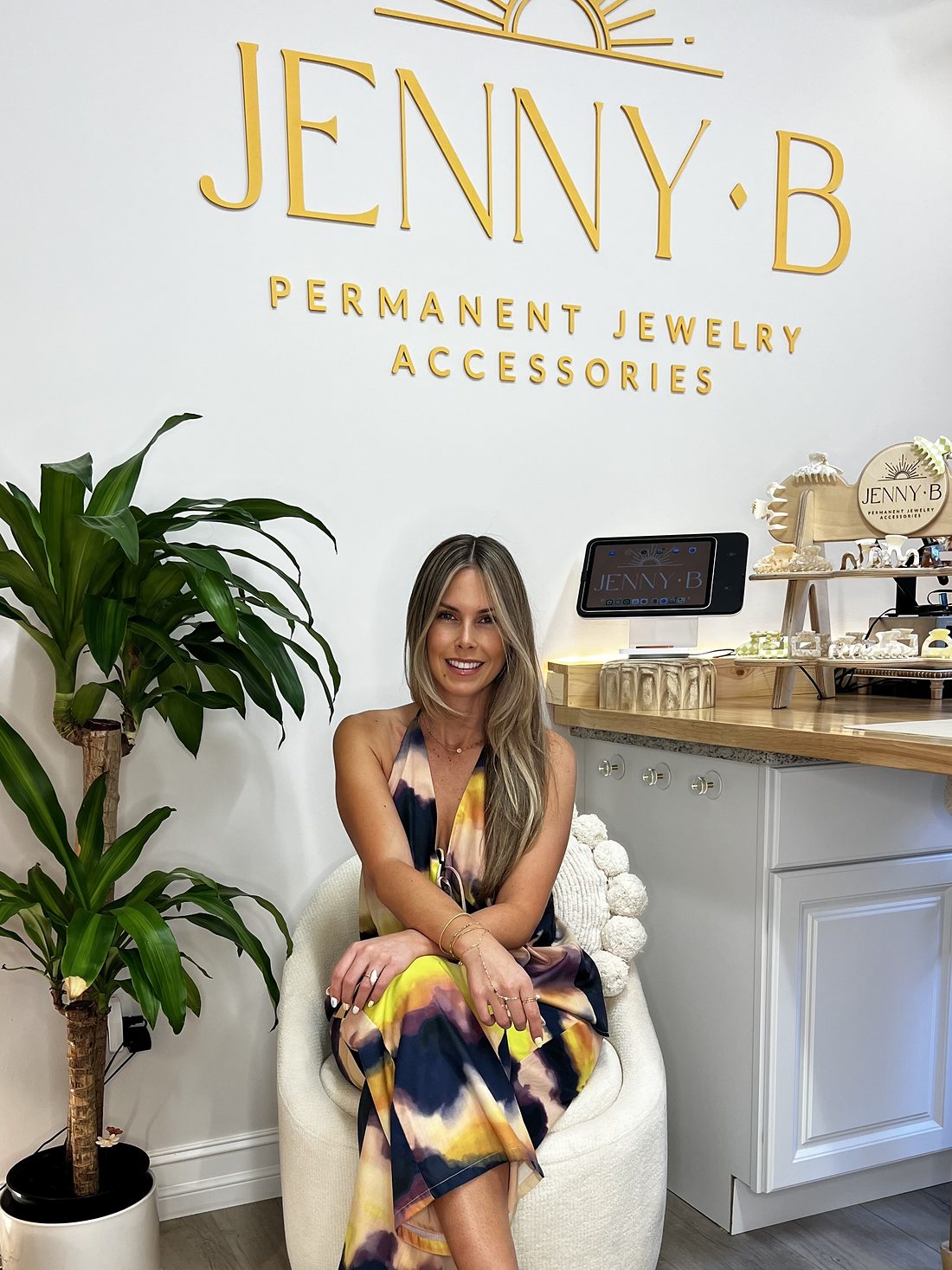 Jenny, owner of Jenny B, smiling in a white chair in her Naples studio beneath the gold Jenny B logo.
